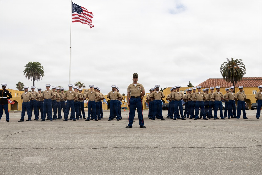 A formal U.S. Marine Corps formation is assembled on a parade deck, with a drill instructor standing at the front center facing the camera. The Marines are dressed in Service Charlie uniform (khaki shirt, blue trousers, white cover) with one figure on the far left in dress blues. An American flag flies prominently on a flagpole in the background, with yellow Spanish Colonial Revival-style buildings and palm trees visible.