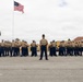 A formal U.S. Marine Corps formation is assembled on a parade deck, with a drill instructor standing at the front center facing the camera. The Marines are dressed in Service Charlie uniform (khaki shirt, blue trousers, white cover) with one figure on the far left in dress blues. An American flag flies prominently on a flagpole in the background, with yellow Spanish Colonial Revival-style buildings and palm trees visible.