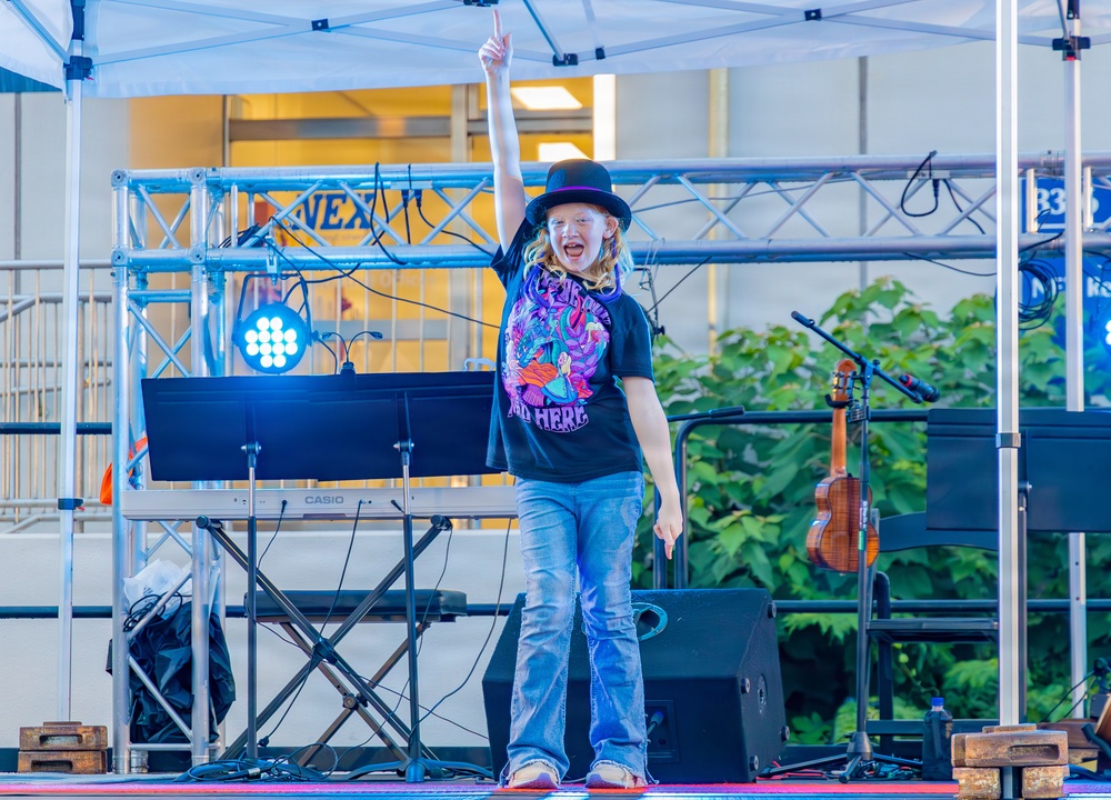 A young performer stands on an outdoor stage under a white canopy tent, raising one finger in the air with an enthusiastic expression. The child is wearing a black graphic t-shirt, jeans, and a black bowler hat, suggesting a performance or talent show setting. Stage equipment including a Casio keyboard, lighting rig, microphone stands, and a ukulele are visible in the background.