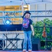 A young performer stands on an outdoor stage under a white canopy tent, raising one finger in the air with an enthusiastic expression. The child is wearing a black graphic t-shirt, jeans, and a black bowler hat, suggesting a performance or talent show setting. Stage equipment including a Casio keyboard, lighting rig, microphone stands, and a ukulele are visible in the background.