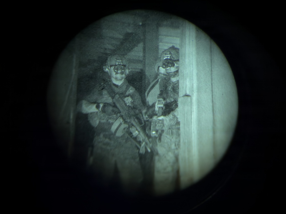 A night-vision or thermal optic view showing two heavily equipped tactical personnel positioned near a doorway or entry point. Both individuals are wearing full tactical gear including helmets with mounted devices, body armor, and carrying firearms. The circular vignette is characteristic of viewing through a night-vision or spotting scope device.