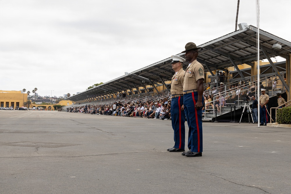 Two U.S. Marines in dress blue uniforms stand at attention on a parade deck in front of a covered grandstand filled with civilian spectators. The setting appears to be a formal military ceremony, likely a graduation or pass-in-review event at a Marine Corps installation. The overcast sky and palm trees in the background suggest a Southern California location, consistent with MCRD San Diego.