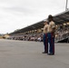 Two U.S. Marines in dress blue uniforms stand at attention on a parade deck in front of a covered grandstand filled with civilian spectators. The setting appears to be a formal military ceremony, likely a graduation or pass-in-review event at a Marine Corps installation. The overcast sky and palm trees in the background suggest a Southern California location, consistent with MCRD San Diego.