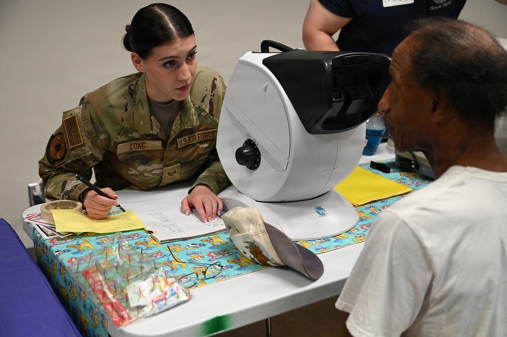 A U.S. Air Force service member named 'Long' in OCP camouflage uniform is conducting what appears to be a medical or vision screening for a civilian patient. The interaction takes place at a table equipped with an autorefractor or similar ophthalmic device, suggesting a community health outreach or humanitarian assistance mission. A second uniformed or staff individual is partially visible in the background.