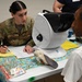 A U.S. Air Force service member named 'Long' in OCP camouflage uniform is conducting what appears to be a medical or vision screening for a civilian patient. The interaction takes place at a table equipped with an autorefractor or similar ophthalmic device, suggesting a community health outreach or humanitarian assistance mission. A second uniformed or staff individual is partially visible in the background.