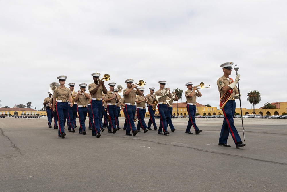 A U.S. Marine Corps marching band is performing on a large paved parade deck, moving in formation from left to right. The musicians are dressed in standard Marine Corps service uniforms with blue trousers and khaki blouses, led by a drum major in a distinctive red-sashed uniform carrying a mace. The setting appears to be a military installation, likely MCRD San Diego, based on the characteristic yellow Spanish Colonial Revival architecture visible in the background.