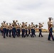 A U.S. Marine Corps marching band is performing on a large paved parade deck, moving in formation from left to right. The musicians are dressed in standard Marine Corps service uniforms with blue trousers and khaki blouses, led by a drum major in a distinctive red-sashed uniform carrying a mace. The setting appears to be a military installation, likely MCRD San Diego, based on the characteristic yellow Spanish Colonial Revival architecture visible in the background.