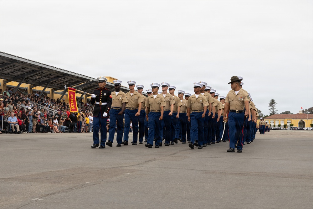 A formation of U.S. Marines in dress uniform is marching in a graduation parade on a large paved drill field. A guidon bearer carries a red and gold unit pennant at the front left of the formation, while an officer leads from the right carrying a sword. A large crowd of spectators watches from bleachers on the left side.