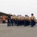 A formation of U.S. Marines in dress uniform is marching in a graduation parade on a large paved drill field. A guidon bearer carries a red and gold unit pennant at the front left of the formation, while an officer leads from the right carrying a sword. A large crowd of spectators watches from bleachers on the left side.