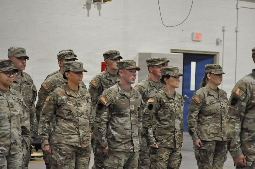 A group of U.S. Army soldiers in Operational Camouflage Pattern (OCP) uniforms are standing in formation inside what appears to be a military armory or indoor facility. Both male and female soldiers are present, wearing standard Army Combat Uniforms with American flag patches on their right shoulders. The setting suggests a formal military ceremony, briefing, or unit assembly.