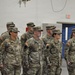 A group of U.S. Army soldiers in Operational Camouflage Pattern (OCP) uniforms are standing in formation inside what appears to be a military armory or indoor facility. Both male and female soldiers are present, wearing standard Army Combat Uniforms with American flag patches on their right shoulders. The setting suggests a formal military ceremony, briefing, or unit assembly.