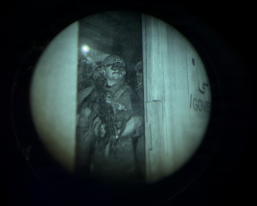 Image captured through a night vision or optical device lens, showing a circular field of view with a military or law enforcement operator visible in a doorway or entry point. The individual is wearing tactical gear including a helmet with night vision device mount and body armor, and appears to be holding a weapon. Partial text visible on a wall to the right reads '/GOVE' suggesting a government building or facility.