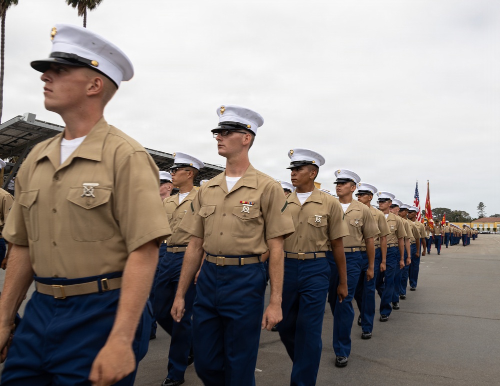 A formation of United States Marines marching in dress uniform during what appears to be a formal parade or graduation ceremony. The Marines are wearing the Charlie Service uniform consisting of khaki short-sleeve shirts, blue trousers, white covers (caps), and gold belts. Color guard personnel carrying the American flag and Marine Corps flag are visible in the background.
