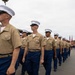 A formation of United States Marines marching in dress uniform during what appears to be a formal parade or graduation ceremony. The Marines are wearing the Charlie Service uniform consisting of khaki short-sleeve shirts, blue trousers, white covers (caps), and gold belts. Color guard personnel carrying the American flag and Marine Corps flag are visible in the background.