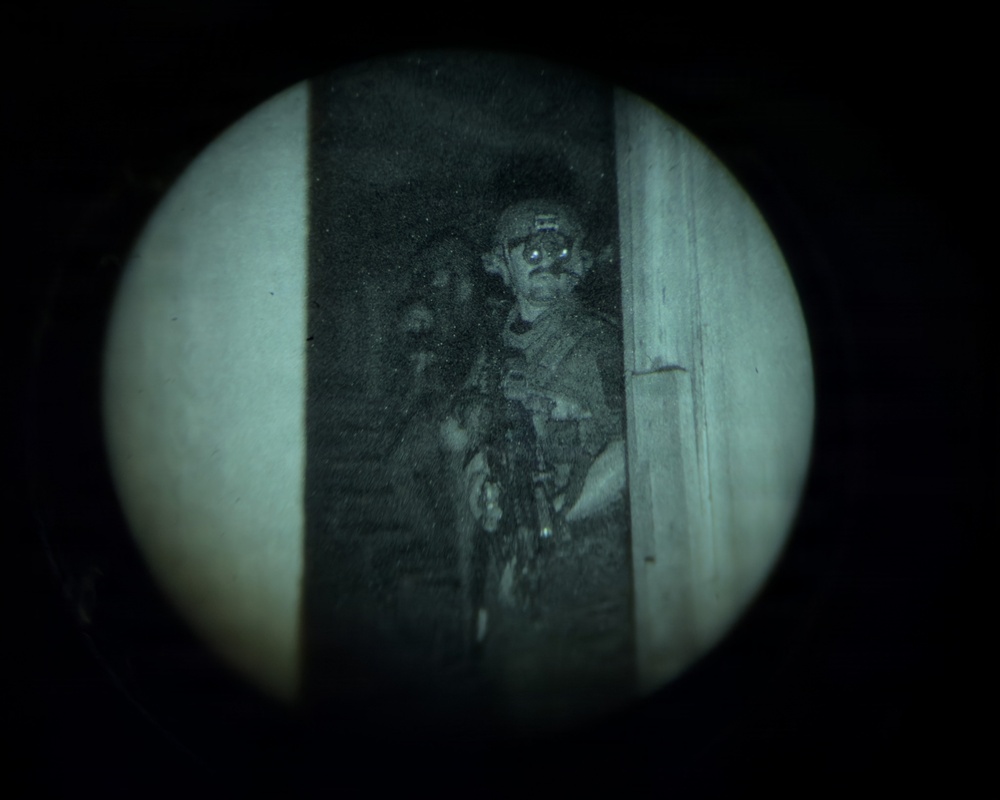 Image captured through a night vision or optical device showing a circular field of view. One or two armed individuals in tactical gear are visible standing in or near a doorway. The image has a characteristic green-gray monochromatic tone typical of low-light optical equipment.