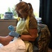 A US Air Force medical personnel member wearing an OCP (Operational Camouflage Pattern) uniform, yellow disposable apron, and blue nitrile gloves is providing wound care or medical treatment to a patient's lower leg/ankle area. The setting appears to be a temporary or field medical station, likely at a community health event or military medical support operation. The provider is wearing an Apple Watch and glasses while focused on the procedure.