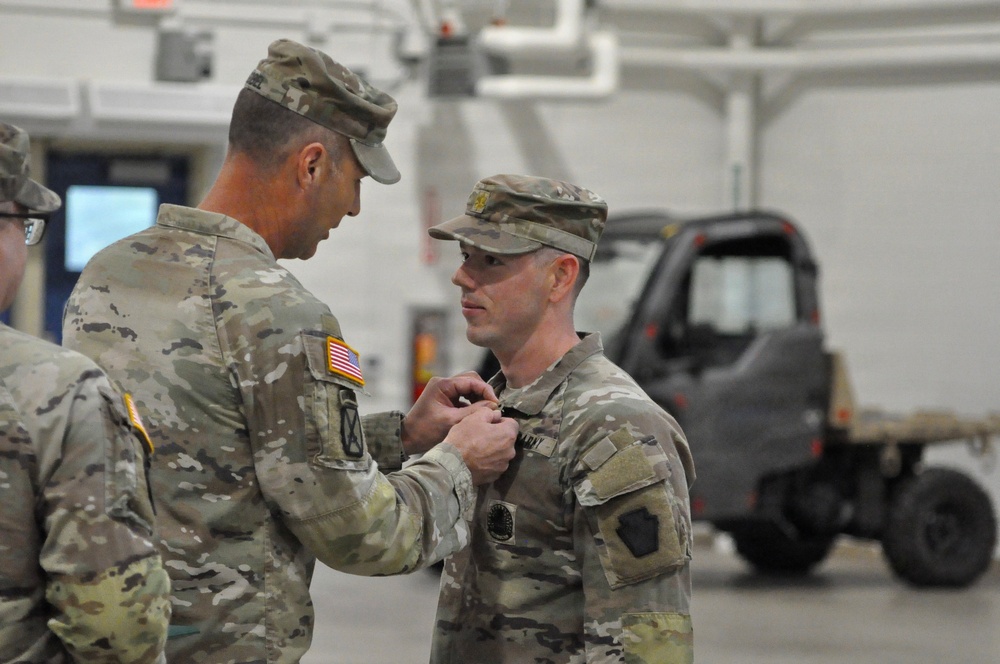 Two U.S. Army soldiers in OCP (Operational Camouflage Pattern) uniforms are engaged in a military decoration or award ceremony inside what appears to be a military hangar or motor pool facility. One soldier is pinning a medal or badge onto the chest of the other soldier, who stands at attention. A third soldier is partially visible on the left side of the frame.