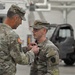 Two U.S. Army soldiers in OCP (Operational Camouflage Pattern) uniforms are engaged in a military decoration or award ceremony inside what appears to be a military hangar or motor pool facility. One soldier is pinning a medal or badge onto the chest of the other soldier, who stands at attention. A third soldier is partially visible on the left side of the frame.