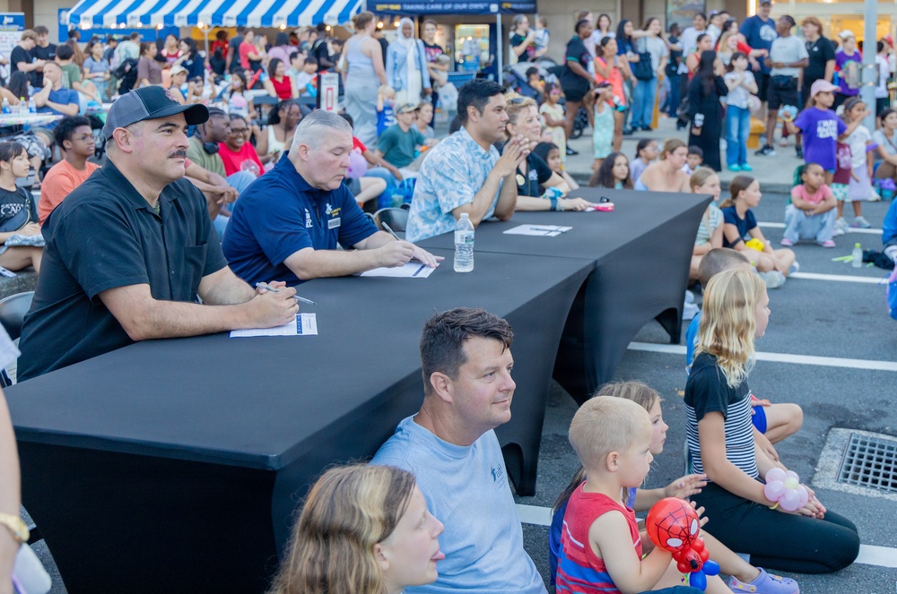 An outdoor community event taking place in a parking lot, featuring a panel of judges or officials seated at a long black-draped table. A large crowd of families, children, and adults are gathered as an audience, sitting on the ground or standing in the background. The setting appears to be a daytime public event near a retail or commercial establishment.