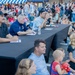 An outdoor community event taking place in a parking lot, featuring a panel of judges or officials seated at a long black-draped table. A large crowd of families, children, and adults are gathered as an audience, sitting on the ground or standing in the background. The setting appears to be a daytime public event near a retail or commercial establishment.