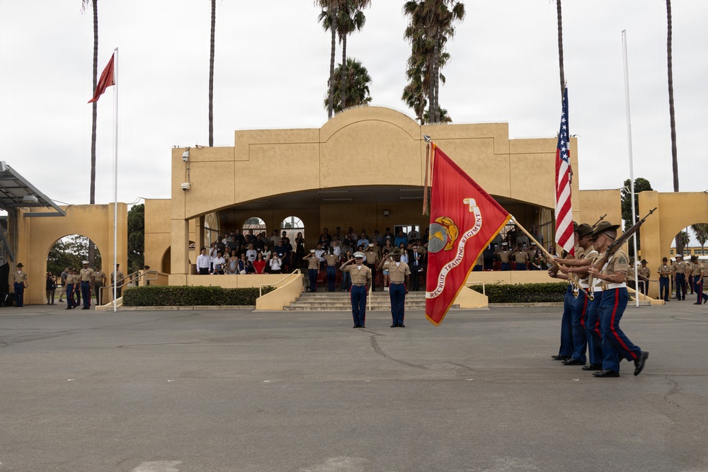 A United States Marine Corps color guard ceremony is taking place on a parade deck, with Marines carrying the U.S. flag and a red 'Recruit Training Regiment' guidon. Two Marines in dress blues stand at salute in the center foreground, while an audience of civilians and military personnel observes from the steps and covered pavilion of a tan stucco building. The setting appears to be MCRD (Marine Corps Recruit Depot) San Diego based on the distinctive architecture and regiment flag.