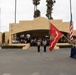 A United States Marine Corps color guard ceremony is taking place on a parade deck, with Marines carrying the U.S. flag and a red 'Recruit Training Regiment' guidon. Two Marines in dress blues stand at salute in the center foreground, while an audience of civilians and military personnel observes from the steps and covered pavilion of a tan stucco building. The setting appears to be MCRD (Marine Corps Recruit Depot) San Diego based on the distinctive architecture and regiment flag.