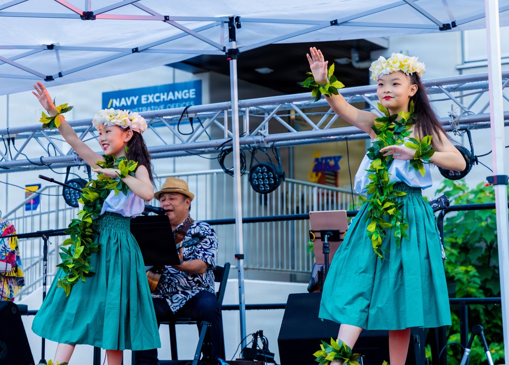 Two young girls in traditional Hawaiian hula costumes perform on an outdoor stage, wearing green ti-leaf leis and white plumeria flower head crowns with teal skirts. A male musician sits behind them playing an instrument and singing into a microphone. The performance is taking place at a Navy Exchange facility, likely at a military base.