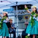 Two young girls in traditional Hawaiian hula costumes perform on an outdoor stage, wearing green ti-leaf leis and white plumeria flower head crowns with teal skirts. A male musician sits behind them playing an instrument and singing into a microphone. The performance is taking place at a Navy Exchange facility, likely at a military base.