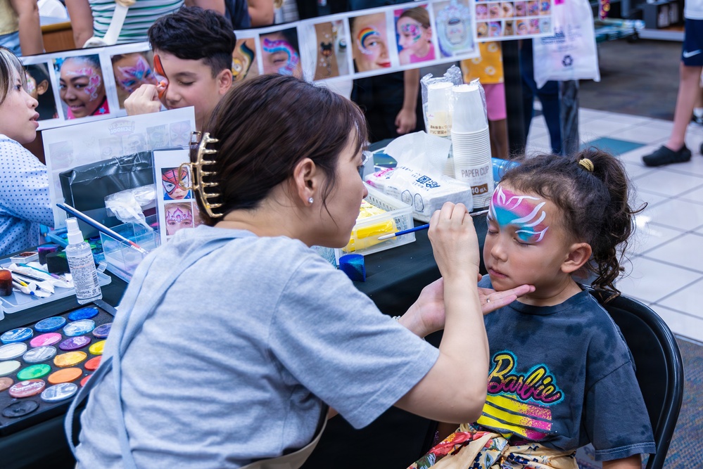 A face painter is applying colorful artistic face paint to a young girl at what appears to be a public event or fair. The girl, wearing a Barbie t-shirt, has a partially completed pink, blue, and white design around her eye area. A display banner showing various face painting examples is visible in the background along with painting supplies on the table.