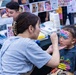 A face painter is applying colorful artistic face paint to a young girl at what appears to be a public event or fair. The girl, wearing a Barbie t-shirt, has a partially completed pink, blue, and white design around her eye area. A display banner showing various face painting examples is visible in the background along with painting supplies on the table.
