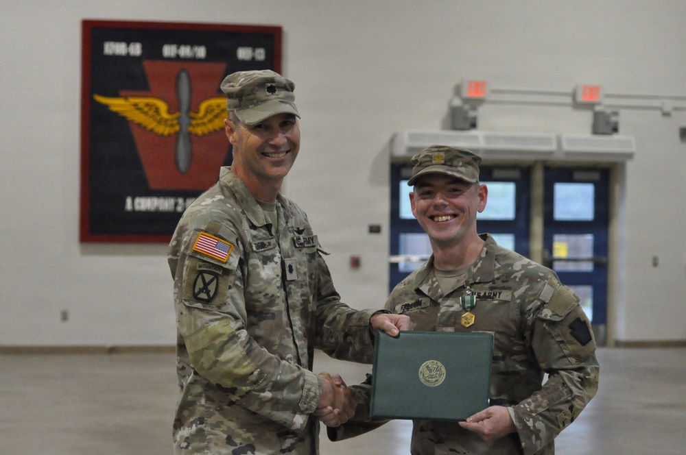 Two U.S. Army soldiers are posing together during what appears to be a military awards ceremony. The soldier on the right is receiving a certificate and is wearing a medal on his chest, while the soldier on the left, who has a lieutenant colonel rank insignia on his cap, is presenting the award with a handshake. The ceremony is taking place inside a military facility with a unit patch/insignia displayed on the wall behind them.