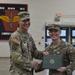 Two U.S. Army soldiers are posing together during what appears to be a military awards ceremony. The soldier on the right is receiving a certificate and is wearing a medal on his chest, while the soldier on the left, who has a lieutenant colonel rank insignia on his cap, is presenting the award with a handshake. The ceremony is taking place inside a military facility with a unit patch/insignia displayed on the wall behind them.