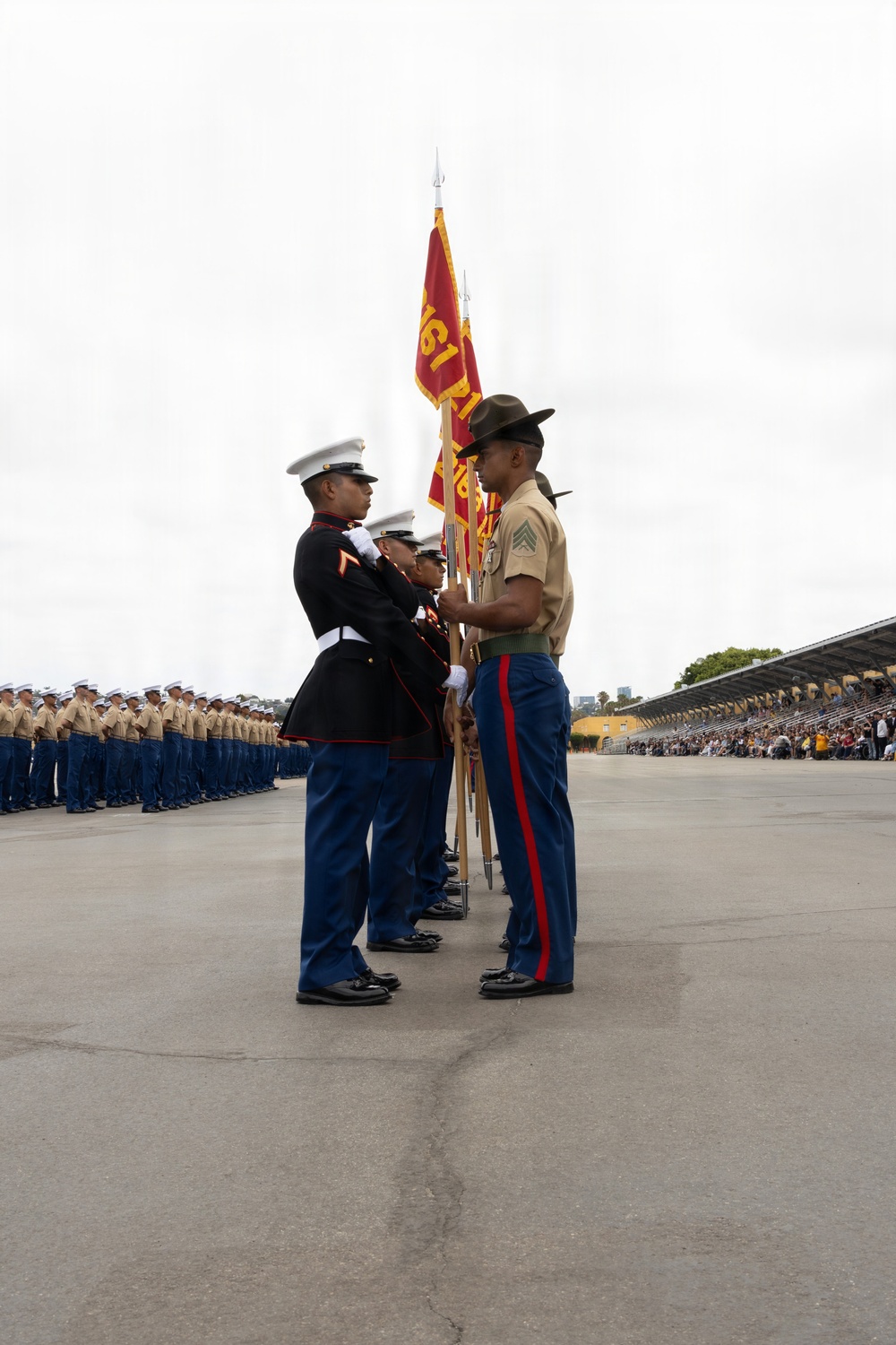 A U.S. Marine Corps graduation ceremony (likely a recruit training graduation) is taking place on a parade deck. A drill instructor in a campaign cover and service uniform is presenting guidon flags to Marines in dress blue uniforms. Rows of newly graduated Marines in service uniforms and a grandstand with spectators are visible in the background.