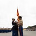 A U.S. Marine Corps graduation ceremony (likely a recruit training graduation) is taking place on a parade deck. A drill instructor in a campaign cover and service uniform is presenting guidon flags to Marines in dress blue uniforms. Rows of newly graduated Marines in service uniforms and a grandstand with spectators are visible in the background.