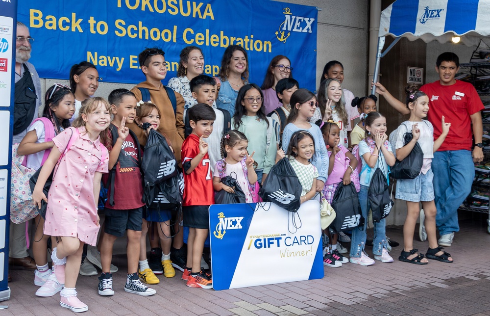 A group of children and adults pose together at a 'Yokosuka Back to School Celebration' hosted by the Navy Exchange (NEX) in Japan. Several children are holding New Balance drawstring backpacks, and one child is holding a large NEX Gift Card Winner sign. The event appears to be a school supply giveaway or contest celebration at a military base exchange.
