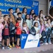 A group of children and adults pose together at a 'Yokosuka Back to School Celebration' hosted by the Navy Exchange (NEX) in Japan. Several children are holding New Balance drawstring backpacks, and one child is holding a large NEX Gift Card Winner sign. The event appears to be a school supply giveaway or contest celebration at a military base exchange.