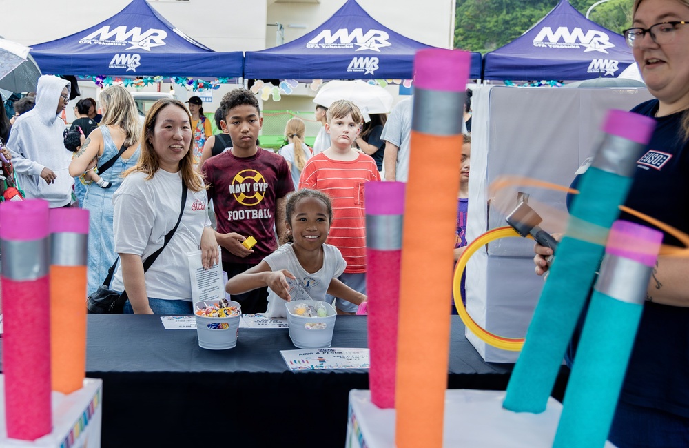 A family-friendly outdoor event organized by MWR (Morale, Welfare, and Recreation) at CFА Yokosuka, a U.S. Navy installation in Japan. Participants are gathered around a game booth featuring a ring-toss activity using oversized pencil-shaped posts. Children and adults are engaging with the activity booth while staff assist.