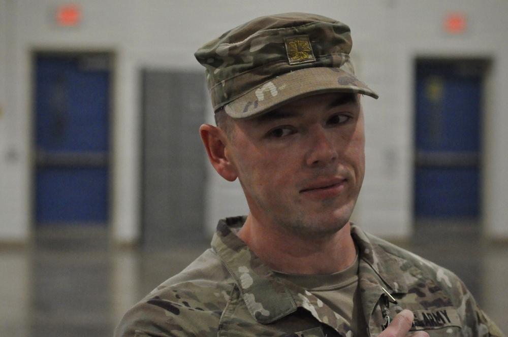 A young male U.S. Army soldier is shown in close-up, wearing Operational Camouflage Pattern (OCP) uniform and patrol cap with a unit crest insignia. He appears to be indoors in a military facility, possibly speaking or being interviewed. The background shows blue-doored lockers or storage units in a blurred gymnasium or armory-type space.