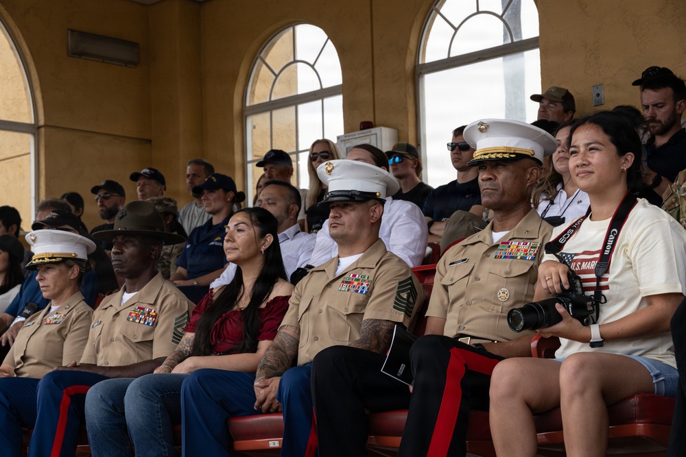 A group of U.S. Marine Corps personnel in service uniforms (tan/khaki) are seated in stadium-style bleachers alongside civilian companions, appearing to observe a formal event or ceremony. Several Marines wear distinctive white dress covers (caps) indicating senior or officer rank, and display multiple service ribbons and medals. The setting appears to be an indoor or covered grandstand venue with arched windows.