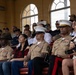 A group of U.S. Marine Corps personnel in service uniforms (tan/khaki) are seated in stadium-style bleachers alongside civilian companions, appearing to observe a formal event or ceremony. Several Marines wear distinctive white dress covers (caps) indicating senior or officer rank, and display multiple service ribbons and medals. The setting appears to be an indoor or covered grandstand venue with arched windows.