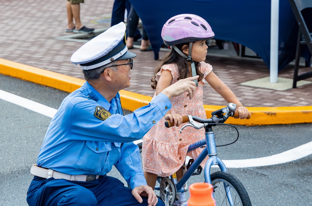 A uniformed officer, appearing to be Japanese police based on the badge and uniform style, kneels beside a young girl seated on a small blue bicycle. The officer is pointing and providing instruction, likely as part of a bicycle safety or traffic education event for children. The setting appears to be an outdoor traffic safety training area marked with white lines and yellow curbing.