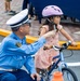 A uniformed officer, appearing to be Japanese police based on the badge and uniform style, kneels beside a young girl seated on a small blue bicycle. The officer is pointing and providing instruction, likely as part of a bicycle safety or traffic education event for children. The setting appears to be an outdoor traffic safety training area marked with white lines and yellow curbing.