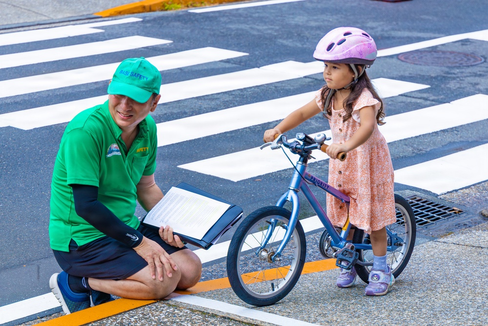 A safety instructor wearing a green 'RASI SAFETY' shirt and cap crouches beside a young girl on a small blue 'Priority' brand bicycle at a crosswalk. The child is wearing a pink helmet and a floral dress, appearing to be participating in a bicycle safety or skills assessment. The setting is an urban street intersection with clearly marked crosswalk stripes and a storm drain visible.