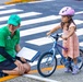 A safety instructor wearing a green 'RASI SAFETY' shirt and cap crouches beside a young girl on a small blue 'Priority' brand bicycle at a crosswalk. The child is wearing a pink helmet and a floral dress, appearing to be participating in a bicycle safety or skills assessment. The setting is an urban street intersection with clearly marked crosswalk stripes and a storm drain visible.