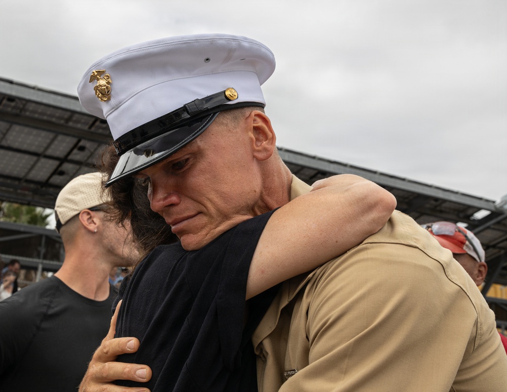 A U.S. Marine in dress uniform shares an emotional embrace with a civilian, likely during a homecoming or farewell event. The Marine's expression conveys deep emotion, with eyes closed and a visibly moved expression. The setting appears to be an outdoor gathering with other people visible in the background.