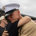 A U.S. Marine in dress uniform shares an emotional embrace with a civilian, likely during a homecoming or farewell event. The Marine's expression conveys deep emotion, with eyes closed and a visibly moved expression. The setting appears to be an outdoor gathering with other people visible in the background.