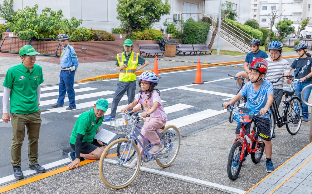 A bicycle safety training event for children is taking place in what appears to be a Japanese urban setting. Instructors wearing green 'Safety' shirts and a police officer are supervising children on bicycles navigating a marked crosswalk area. The event appears to be an organized traffic safety education program with cones, road markings, and safety equipment in use.