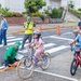 A bicycle safety training event for children is taking place in what appears to be a Japanese urban setting. Instructors wearing green 'Safety' shirts and a police officer are supervising children on bicycles navigating a marked crosswalk area. The event appears to be an organized traffic safety education program with cones, road markings, and safety equipment in use.