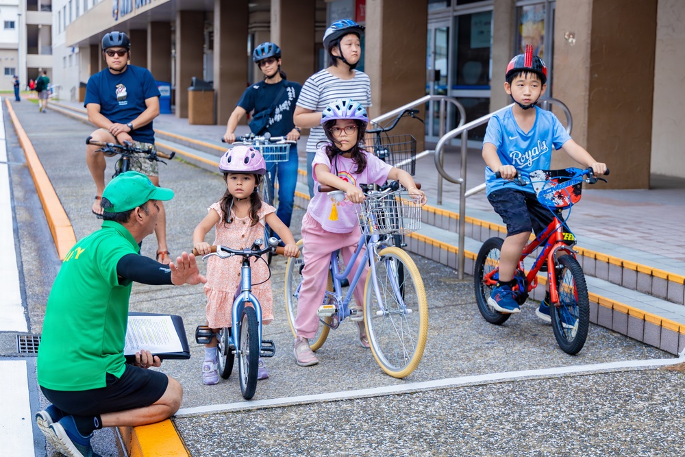 A group of children on bicycles are receiving instruction from an adult in a green shirt and cap, who is crouching down and gesturing toward them. The scene appears to be a structured cycling lesson or safety event taking place in an outdoor urban area, likely near a school or community building. Two adult supervisors on bicycles are visible in the background.
