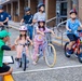 A group of children on bicycles are receiving instruction from an adult in a green shirt and cap, who is crouching down and gesturing toward them. The scene appears to be a structured cycling lesson or safety event taking place in an outdoor urban area, likely near a school or community building. Two adult supervisors on bicycles are visible in the background.